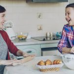Young mother rolling dough and talking to her little cute daughter while cooking in the kitchen on weekend. Family, food, home and people concept